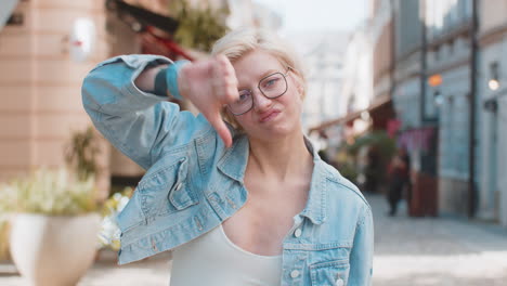 upset young caucasian blonde woman showing double thumbs down expressing discontent on city street