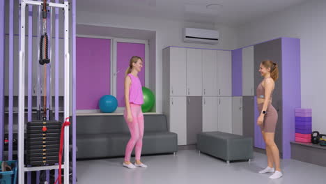 instructor en ropa deportiva marrón da la bienvenida a un amigo en rosa con una cálida sonrisa en un gimnasio moderno con decoración pastel, casilleros, equipos de ejercicio, máquina de resistencia, bolas de fitness y una cómoda zona de asientos