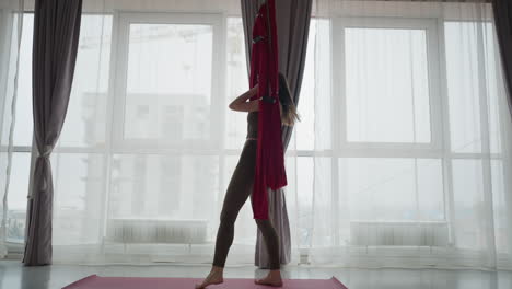 low angle view of woman in olive activewear climbing red silk hammock above yoga mat on floor in bright studio with sheer curtains and windows showing urban house view, conveying balance strength