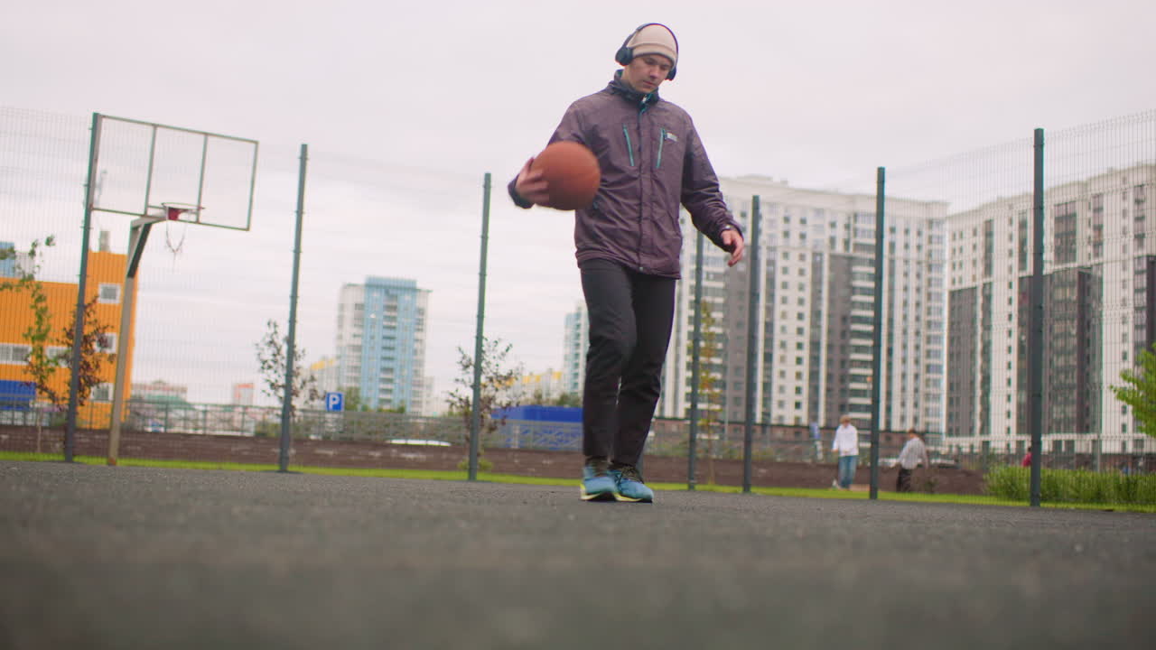 uomo in un campo da basket urbano che palleggia all'aperto, con felpa e scarpe da ginnastica, torri di appartamenti che si ergono sullo sfondo sotto un cielo nuvoloso, primo piano ad angolo basso sul rimbalzo della palla e sui piedi, sessione di allenamento individuale incentrata