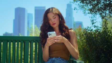 Calm-businesswoman-scrolling-phone-in-park.-Asian-lady-sitting-on-bench-outdoors