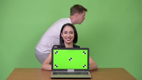 Young beautiful Asian businesswoman showing laptop with young man passing by