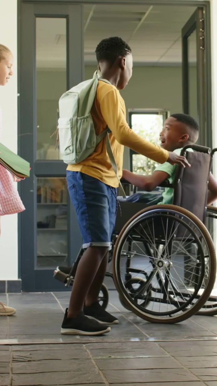 Video verticale: Entrando dalla porta della scuola, un bambino in sedia a rotelle che ride mentre un assistente lo guida in classe.