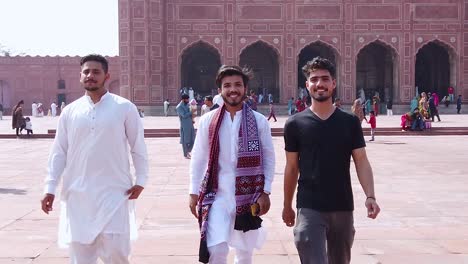 slow motion backward dolly shot of three pakistani boys walking and smiling at the camera with badshahi mosque in the back, lahore pakistan