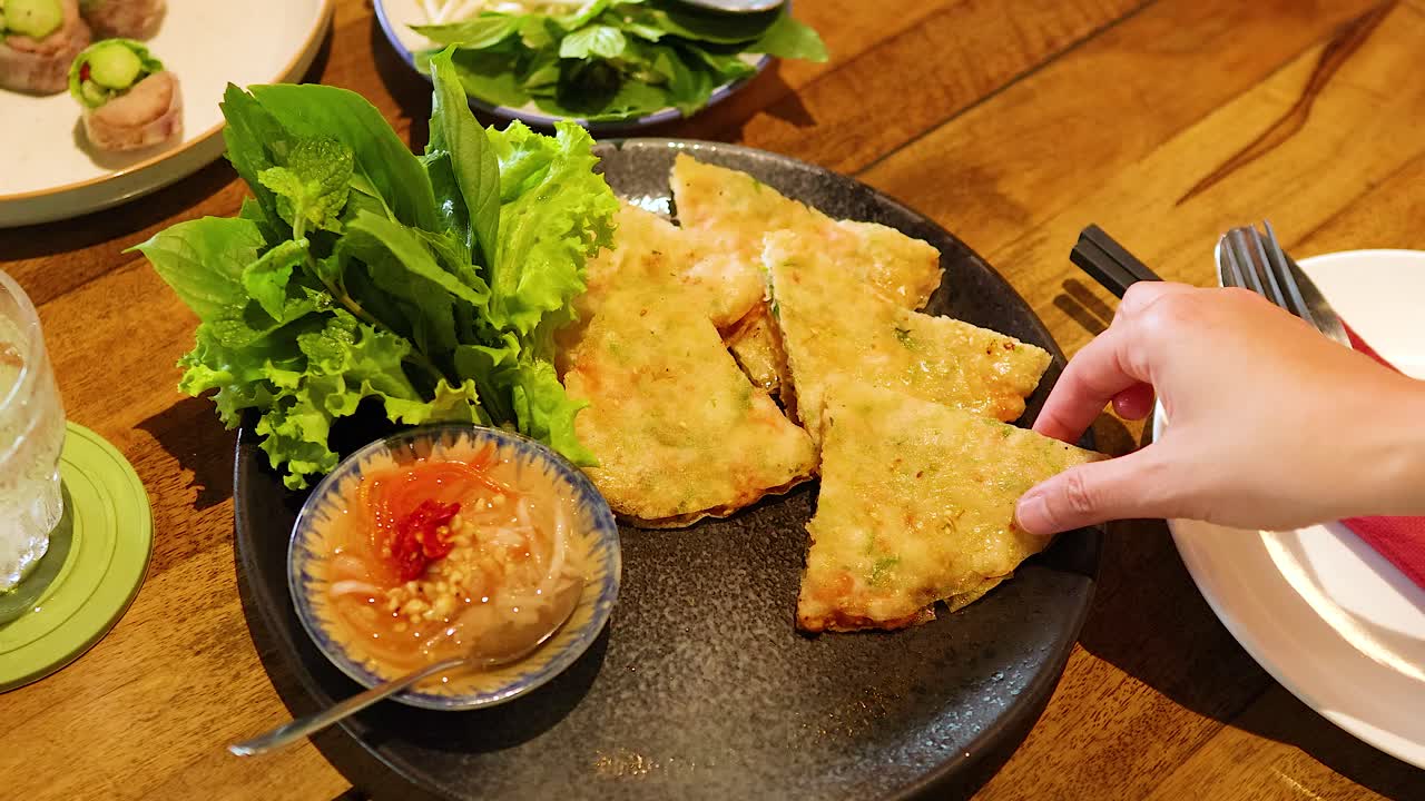 Hands reach for crispy shrimp pancakes with greens and sauce on a wooden table. Bright lighting enhances the vibrant colors