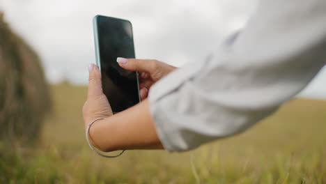 close-up hand view of lady in long sleeve taking photo of someone in the background, tall grass surrounds her as she focuses on capturing the moment, with a softly blurred countryside landscape