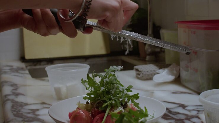 A detailed close-up shot featuring a person's hands using a microplane to grate fresh cheese or zest over a vibrant salad of arugula and cherry tomatoes on a white marble countertop