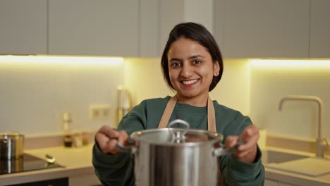 portrait of a happy brunette indian girl in a green jacket and a beige apron holding a gray shiny pan in her hands and looking at the camera in a modern kitchen