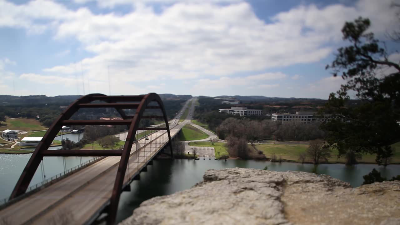 Austin Pennybacker Bridge tilt-shifted time-lapse, focus isolated on center of bridge and boat ramp across the river, mostly cloudy as the clouds move south.