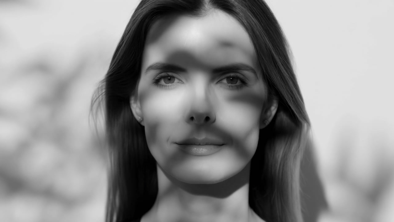Centered monochrome portrait shows a woman looking directly toward camera while patterned shadows drift across her face, ending with a faint smile against a softly blurred background.
