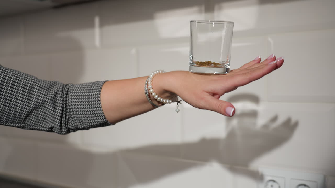 clean glass with beads inside placed on back hand adorned with pearl bracelets, kitchen background with white tiles showcasing delicate hand movement and stylish kitchen deco