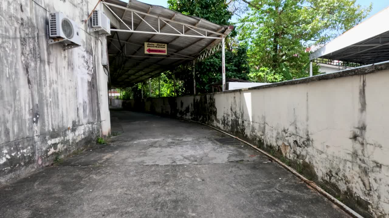 Camera moves along empty alley with old walls, tropical greenery, and covered hotel entrance, daylight