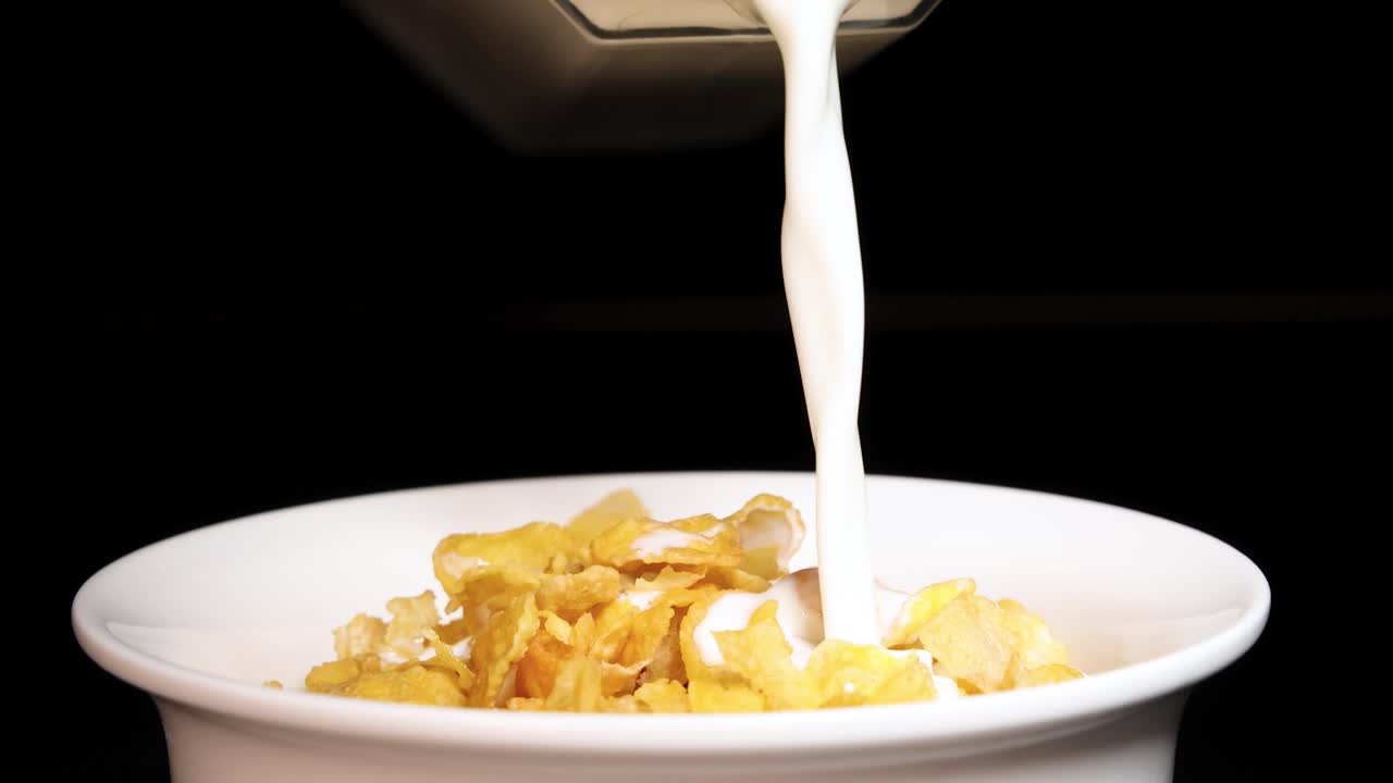 Milk streams into a bowl of crunchy corn cereal against a dark backdrop, highlighting the breakfast preparation