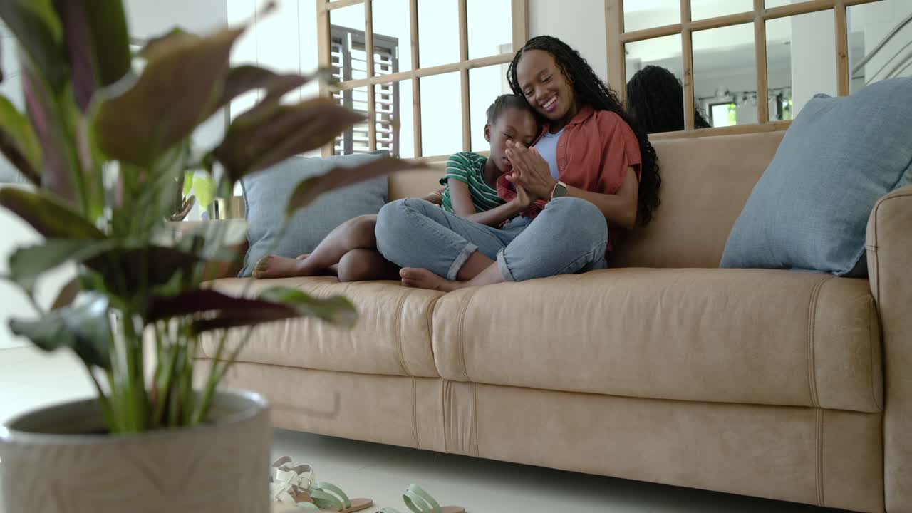 African mother and youth daughter viewing phone, laughing and hugging, checking smartwatch on couch