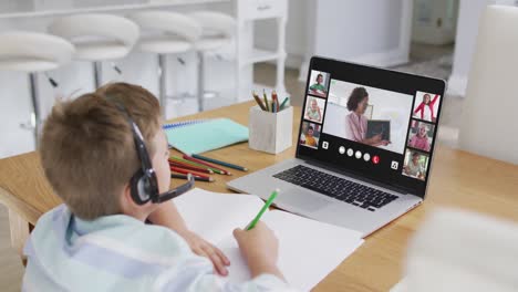 Schoolboy-using-laptop-for-online-lesson-at-home,-with-diverse-teacher-and-class-on-screen