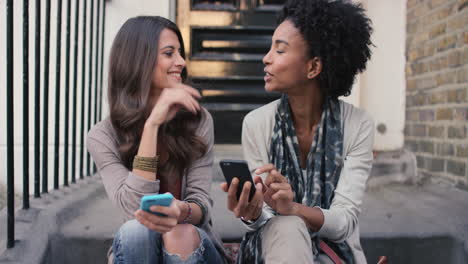 two beautiful woman friends sitting on steps having fun using smart phone