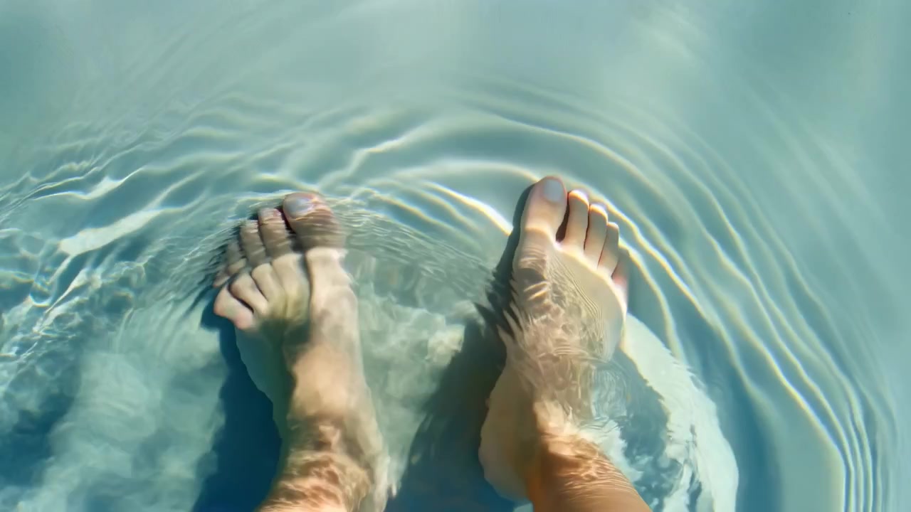 Piedi maschili scalzi che guadano in avanti in una piscina turchese poco profonda, a metà sequenza, sotto la luce solare intensa.