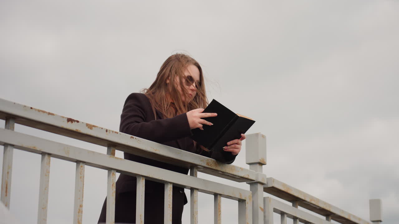 Signora elegante con ciocche castane che indossa un abito lungo legge un libro all'aperto mentre una leggera brezza le muove i capelli, esprimendo eleganza, calma e uno stato d'animo pensieroso sotto un cielo nuvoloso vicino a una ringhiera di metallo