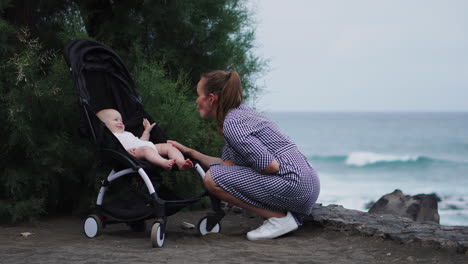 Vor-Der-Ruhigen-Kulisse-Des-Ozeans-Nimmt-Eine-Süße-Junge-Familie-An-Interaktionen-Teil,-Während-Die-Junge-Mutter-Sich-Liebevoll-Um-Ihr-Baby-Im-Kinderwagen-Kümmert-–-Ein-Zeichen-Der-Fürsorge-Und-Zuneigung