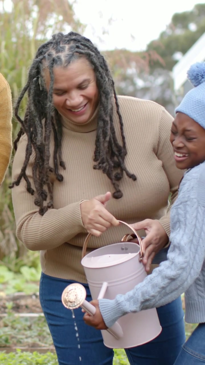 Vertical video: Mother lifting watering can guiding daughter watering seedlings in backyard