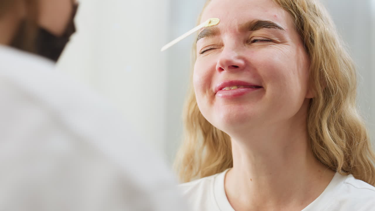 Close up of beautician removing wax from client's eyebrow during facial grooming session. Client smiles with eyes shut while holding still, capturing beauty care moment in bright treatment room