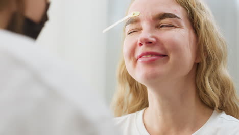 close up of beautician removing wax from client's eyebrow during facial grooming session. client smiles with eyes shut while holding still, capturing beauty care moment in bright treatment room