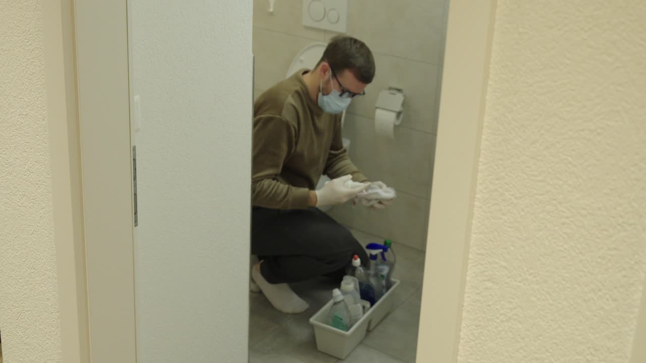 Mann mit Gesichtsmaske und Handschuhen reinigt ein Badezimmer, um Noroviren oder Krankenhauskeime zu verhindern. Konzept von Desinfektion, Hygieneroutine, Infektionskontrolle und Sicherheit in der häuslichen Pflege.