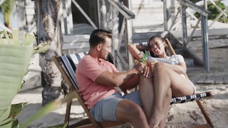 Happy-diverse-couple-sitting-and-drinking-a-toast-with-beers-outside-beach-house,-in-slow-motion
