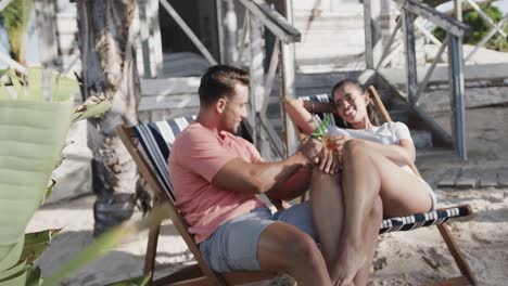 happy diverse couple sitting and drinking a toast with beers outside beach house, in slow motion