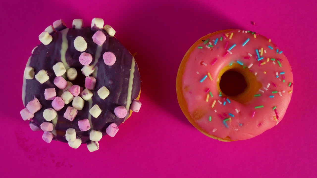 A hand with bright orange nail polish places a pink frosted donut with colorful sprinkles beside a chocolate marshmallow donut on a vivid pink background, creating a playful dessert pairing.