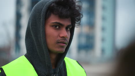 Young male worker in dark hoodie and bright reflective vest standing outdoors in urban area, speaking with serious focused expression, possibly discussing instructions or safety procedures during task