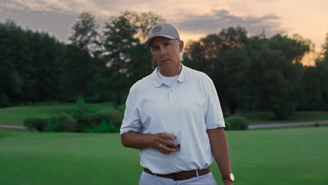 Relaxed-senior-looking-camera-on-green-nature-golf-course.-Old-man-enjoy-drink.