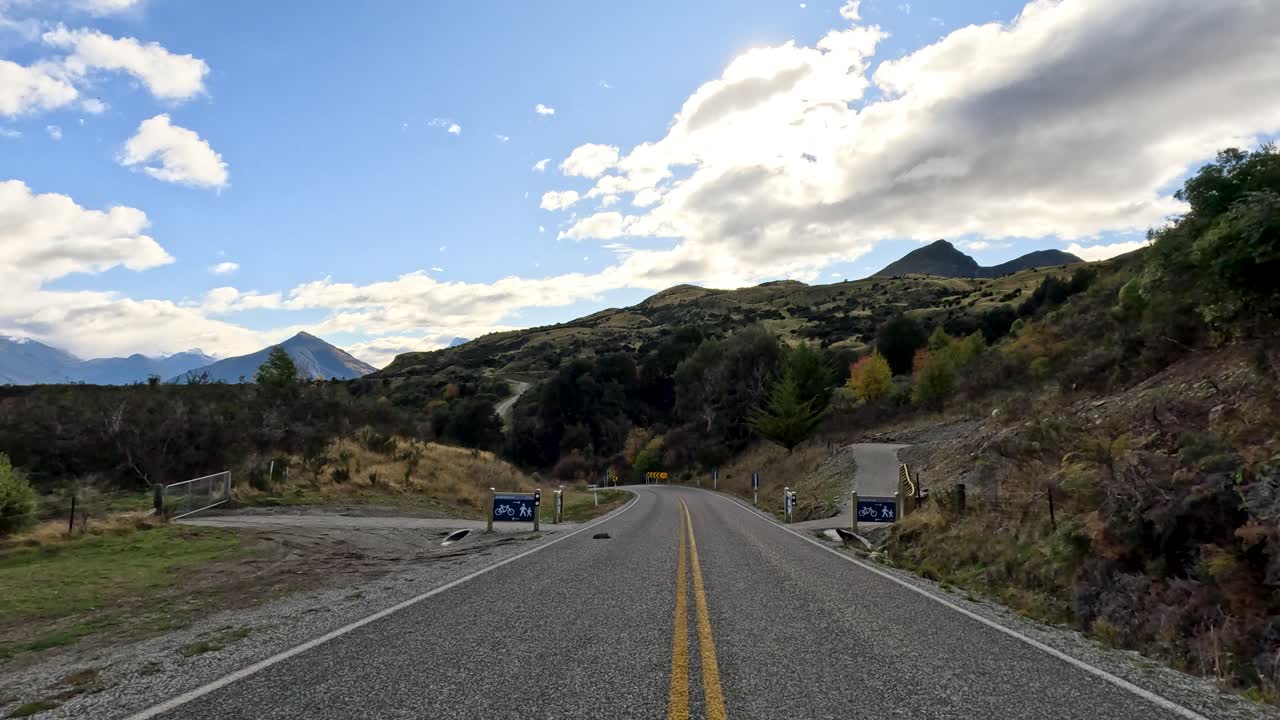 Vehicle travels winding rural road through mountainous landscape, daylight, partly cloudy sky, forward-facing camera