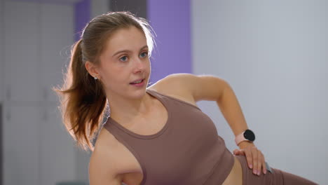 woman in brown sportswear places hand on waist while stretching and conversing, background features well-lit gym with elegant decor, soft lighting, and a bright glow behind her