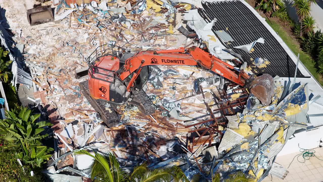 Aerial close-up of an orange excavator demolishing a residential structure amid debris in bright daylight, with steady overhead camera movement and clear shadows
