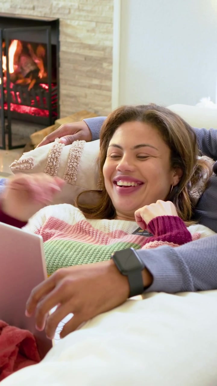 Video verticale: La telecamera si allarga mostrando una donna con maglia a righe che digita sul laptop, mentre un uomo riposa accanto al fuoco.
