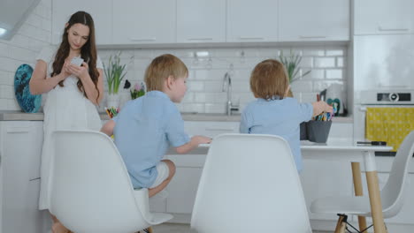 Dos-Hijos-Con-Camisas-Azules-Sentados-En-La-Mesa-Y-Dibujando-Lápices-Sobre-Papel-Sentados-En-Una-Mesa-Blanca,-Y-Mi-Madre-Escribe-Un-Mensaje-De-Texto-En-El-Smartphone.-Mamá-Empresario