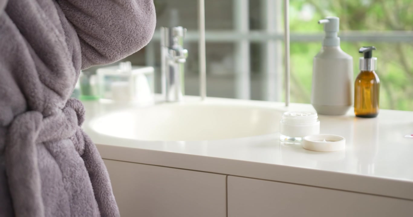 Senior African American woman facing mirror at bathroom sink using cream from jar pump, copy space
