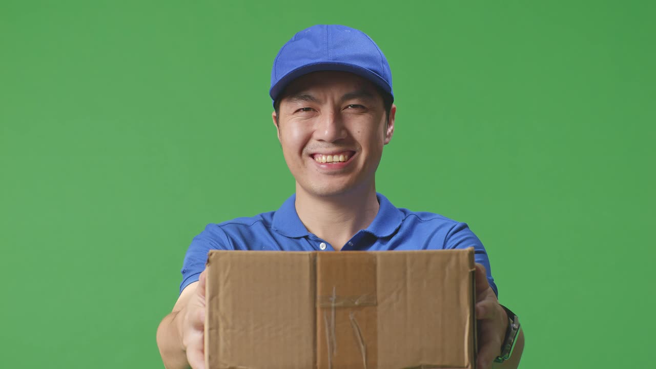 Close Up Of Asian Male Courier In Blue Uniform Showing A Carton To Camera And Smiling While Delivering It On Green Screen Background In The Studio