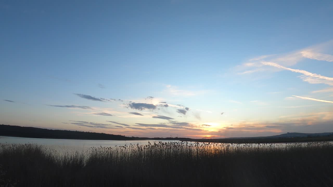 Time lapse at sunset to natural lake. Reeds, clouds, wind, reflections in the water. Smoke in the distance. Plane traces in the sky. Beautiful landscape from Burgas lakes, Bulgaria