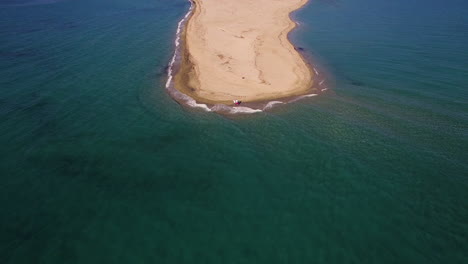 Aerial-View-Two-Travelers-standing-at-the-beach-in-greece-surrounded-by-blue-water