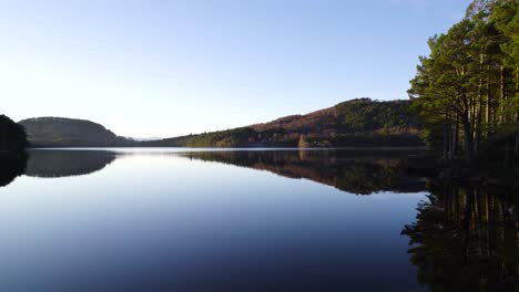 aerial drone footage turning over the surface of still water with reflections in the cairngorms national park scotland towards a native scots pine forest with clear blue sky at sunset