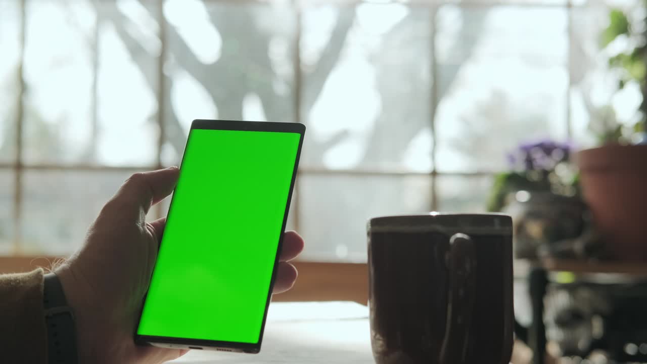 una mano masculina sostiene un teléfono con una pantalla verde de maqueta, y bebe café. disparo de cerca