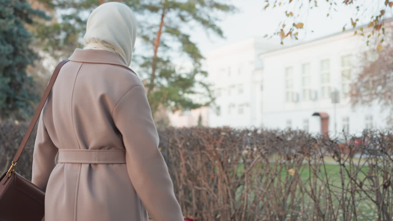 Eine Frau riecht im Herbstpark im Freien an einer roten Rose. Sie trägt einen beigefarbenen Mantel und ein Halstuch. Die nachdenkliche Besucherin atmet den Duft der Blütenblätter ein und legt dann leise eine einzelne Rose auf einen kleinen Gedenkstein neben einer Hecke. Der Himmel ist bedeckt.