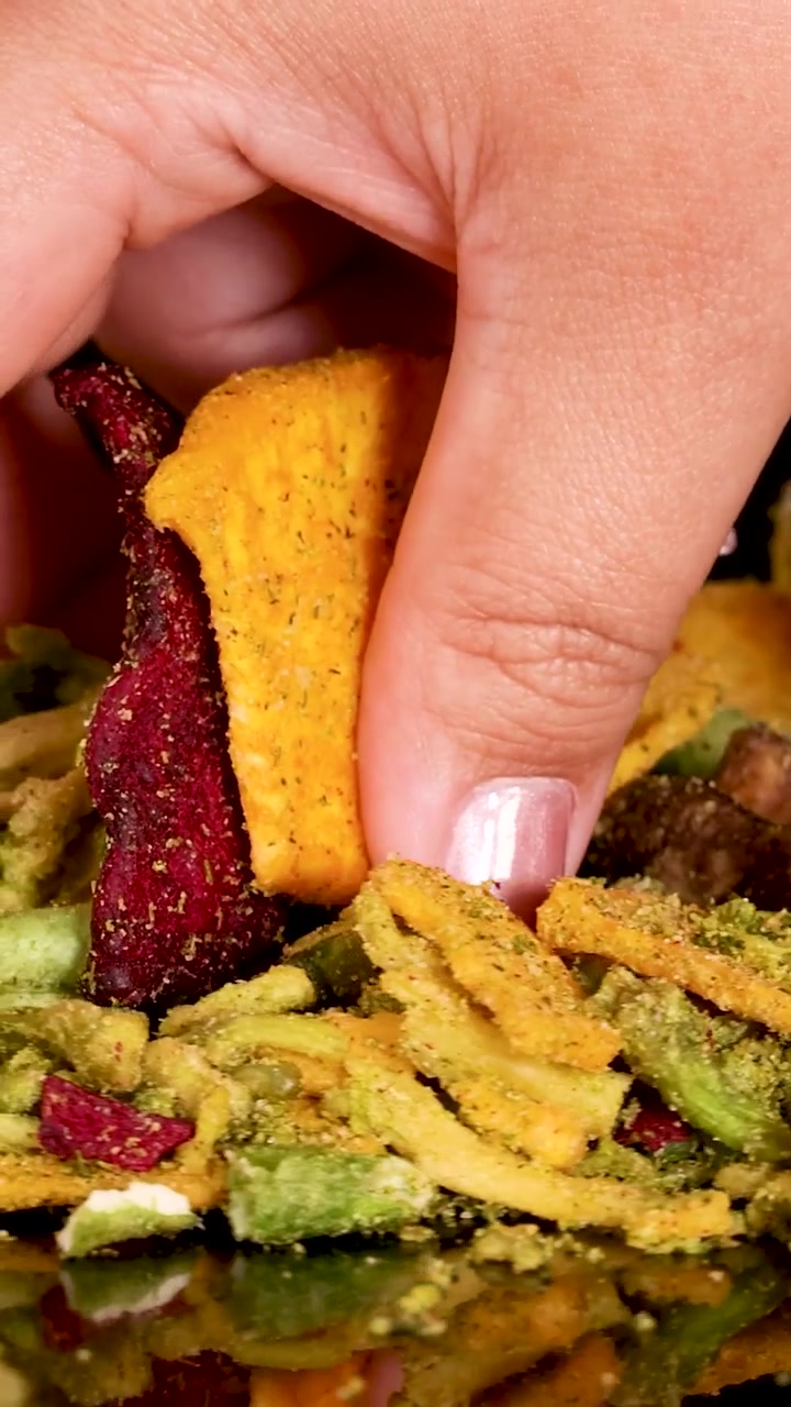 Una mano seleziona chips di verdure essiccate colorate da un mucchio su uno sfondo scuro.