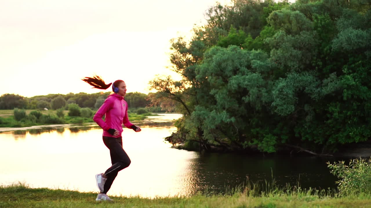 una chica con una chaqueta rosa y pantalones negros corre cerca del río con auriculares preparándose para el maratón