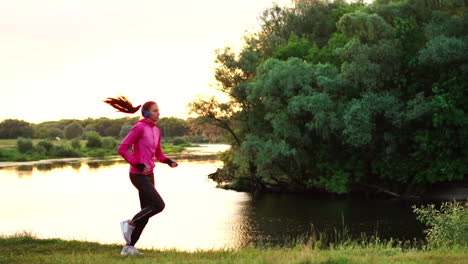 a girl in a pink jacket and black pants runs near the river in headphones preparing for the marathon