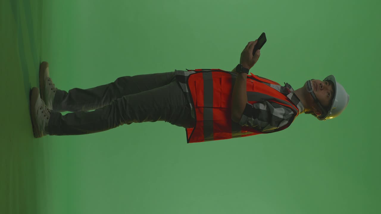 vista lateral completa del cuerpo de un ingeniero masculino asiático con casco de seguridad usando un teléfono inteligente y mirando a su alrededor mientras está de pie en el estudio de fondo de pantalla verde, durante la puesta o el amanecer