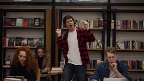 joyful excited handsome european man in checkered shirt performing expressive dance while listening groovy music in headphones in academic library against bookshelves background and classmates around