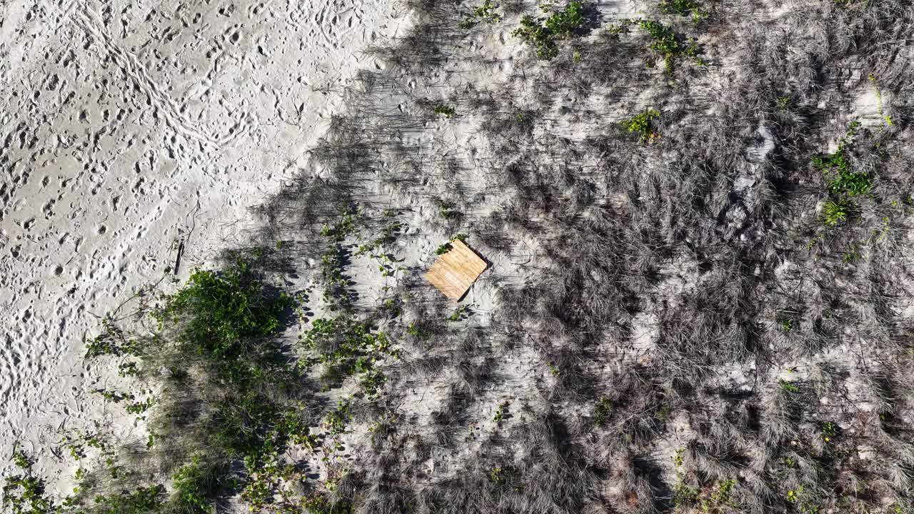 Drone camera smoothly glides above sandy beach and coastal dunes, revealing a small wooden platform amid sparse vegetation in bright daylight